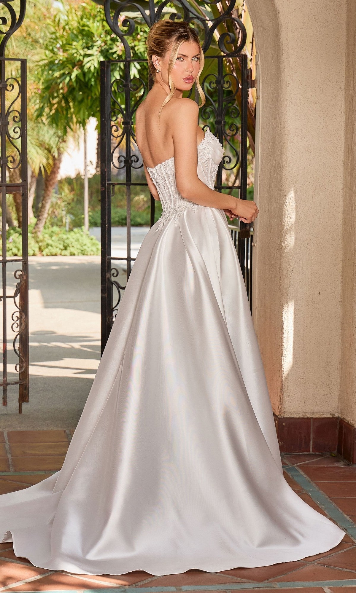 A woman stands near an open wrought iron gate wearing the Ladivine CC511W, a long formal white strapless gown with an A-line silhouette and embroidered corset bodice.
